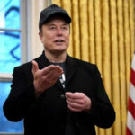 Man wearing hat gesturing in room with flag