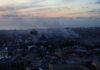 A city skyline at sunset with smoke rising from buildings