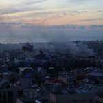A city skyline at sunset with smoke rising from buildings