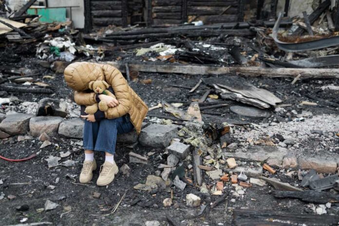 A child sitting on a stone amidst a devastated landscape, holding a teddy bear