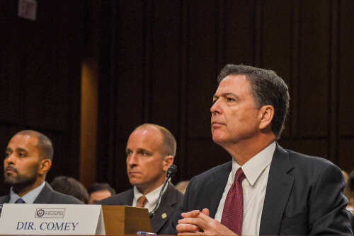 Government officials seated during a hearing with a nameplate in front of one
