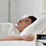 Crescent Pillow REVOLUTION: Does It End Neck Pain? A man sleeping in bed with an alarm clock showing 3:20 AM