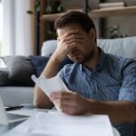 Man sitting in a home office looking stressed while reviewing paperwork