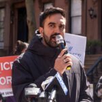 Outrage! NYC Mayor Targets Billionaire on Doorstep Man speaking into a microphone at a protest for senior justice