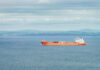 British Raid Rattles Oil — Brace Up Orange cargo ship sailing on calm sea with distant green hills
