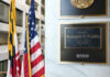 Office plaque beside U.S. and state flags in a government building