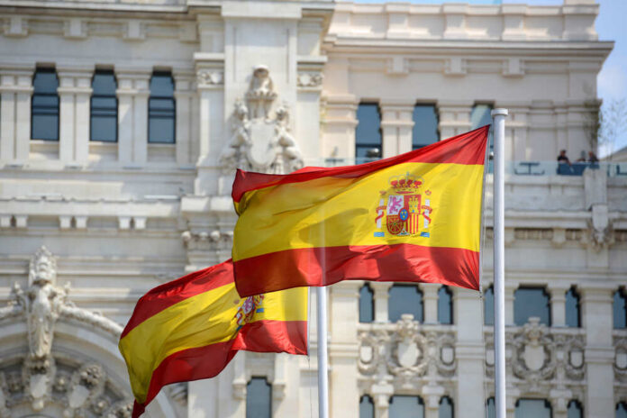 Flag,Of,Spain,In,The,Wind,In,Front,Of,Palace Two Spanish flags waving in front of a historic building