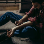 Overdose Survivor Slams America’s Addiction Playbook man sitting on floor injecting his arm with a syringe