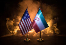 Trump Trades Blocks For Barrels—At What Price? Flags of the United States and Iran displayed on stands with a smoky background