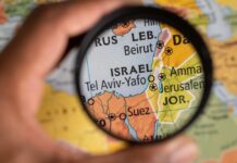 A hand holding a magnifying glass over a map focusing on Israel