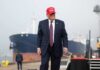 A man in a suit wearing a red cap stands near a ship at a harbor