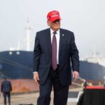 DOJ Power Player Tied To Trump Deal A man in a suit wearing a red cap stands near a ship at a harbor