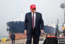 A man in a suit wearing a red cap stands near a ship at a harbor