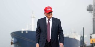 A man in a suit wearing a red cap stands near a ship at a harbor