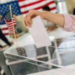 Hand placing ballot in box with American flag nearby