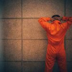 Person in orange jumpsuit stands facing a wall with hands on head