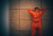 Person in orange jumpsuit stands facing a wall with hands on head
