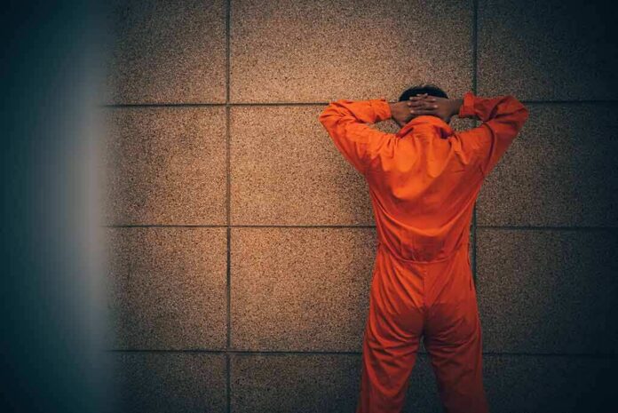 shutterstock_1065732287-1 Person in orange jumpsuit stands facing a wall with hands on head