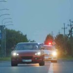 red car driving on road with police car behind using flashing lights