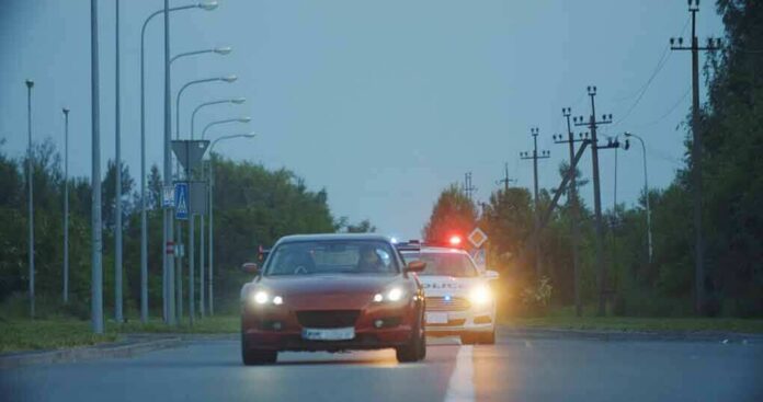 shutterstock_1642255009-3 red car driving on road with police car behind using flashing lights