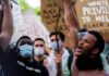 Explosive Claim Collides With DOJ Record Protester shouting with raised fist amid crowd holding signs