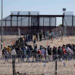 Crowd of people lining up near a border fence with barbed wire