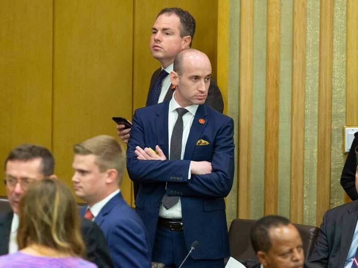 Stephen Miller standing with arms crossed in a formal meeting room