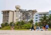 Partially collapsed beachfront apartment building behind people walking on sand