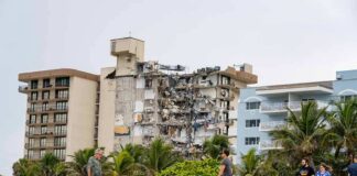 Partially collapsed beachfront apartment building behind people walking on sand