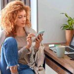 Luxury Couple Dead – $100M Vanished? Woman using smartphone beside laptop on home desk