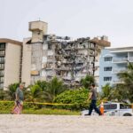Partially collapsed beachfront apartment building behind people walking on sand