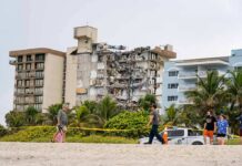 Partially collapsed beachfront apartment building behind people walking on sand