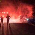 Police officers watch firefighters extinguish a car fire at night